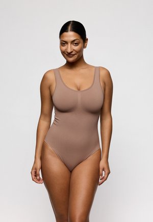 Glunderende vrouw met donker haar in een strakke knot draagt een mouwloos taupe bodypak en staat tegen een effen lichtgrijze achtergrond.