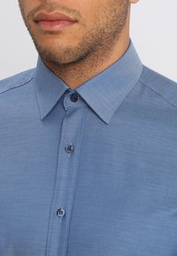 Lichtblauw overhemd met een gestructureerde weving, knoopsluiting aan de kraag en contrasterende knopen, met een subtiel patroon in de stof.