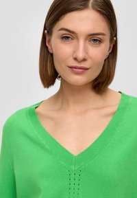 Grüner Strickpullover mit V-Ausschnitt-Design, der subtile Perforationen in der Mitte aufweist, aus weichem Material gefertigt.