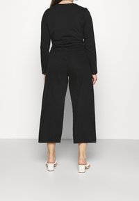 Maglietta nera a maniche lunghe abbinata a pantaloni neri a gamba larga. Il tessuto sembra liscio con un design minimalista. Le scarpe sono décolleté bianchi con tacco squadrato.