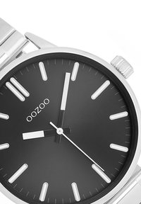 Montre en métal argenté avec un cadran noir, des marqueurs d'heure blancs et des aiguilles contrastantes noir et blanc. Équipée d'un bracelet en acier inoxydable et d'une couronne texturée.