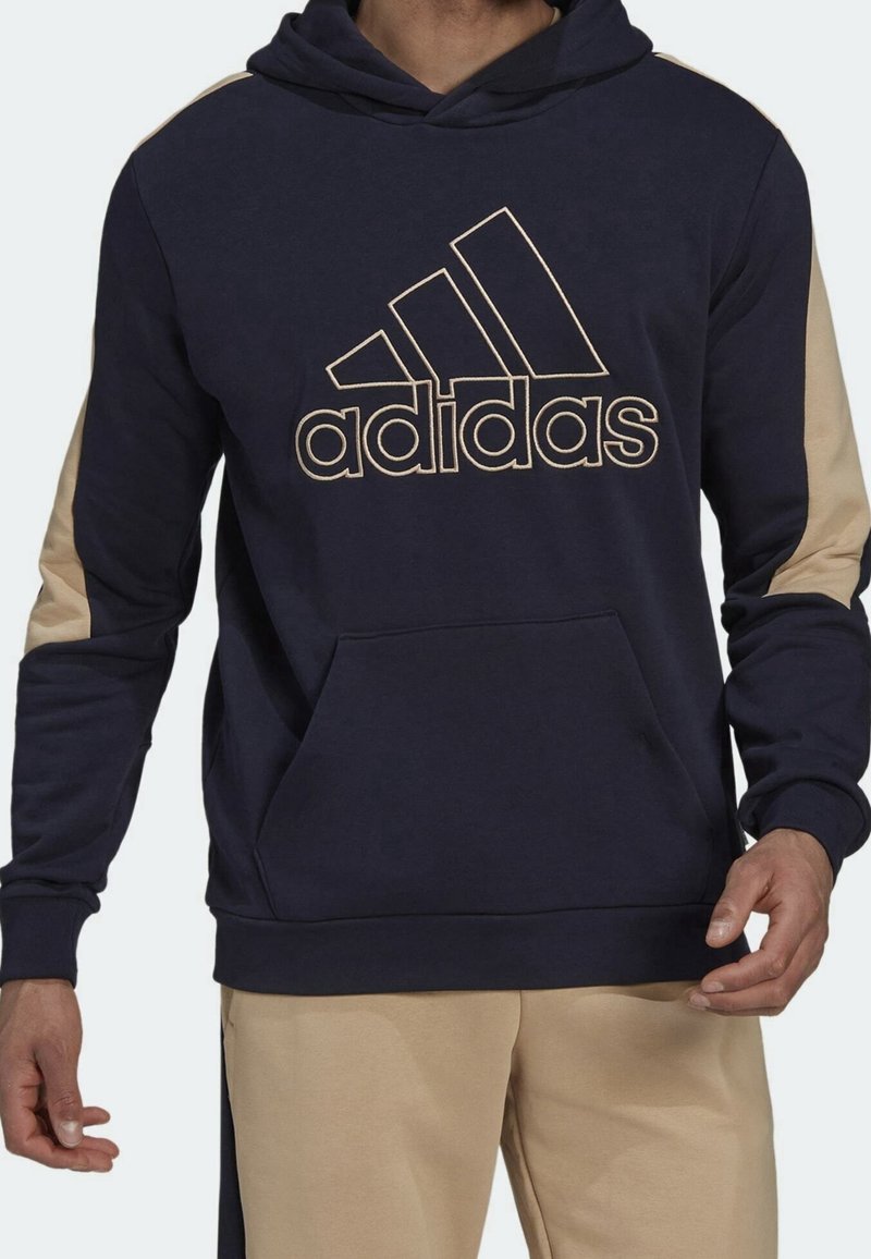 Sweat-shirt à capuche bleu marine avec des accents beiges sur les manches. Présente un grand logo adidas doré et une poche avant. Texture de tissu douce.