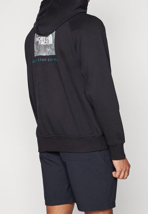 Osoba nosząca czarną bluzę z kapturem North Face z logo oraz napisem „Never Stop Exploring” na plecach, w połączeniu z ciemnoniebieskimi szortami.