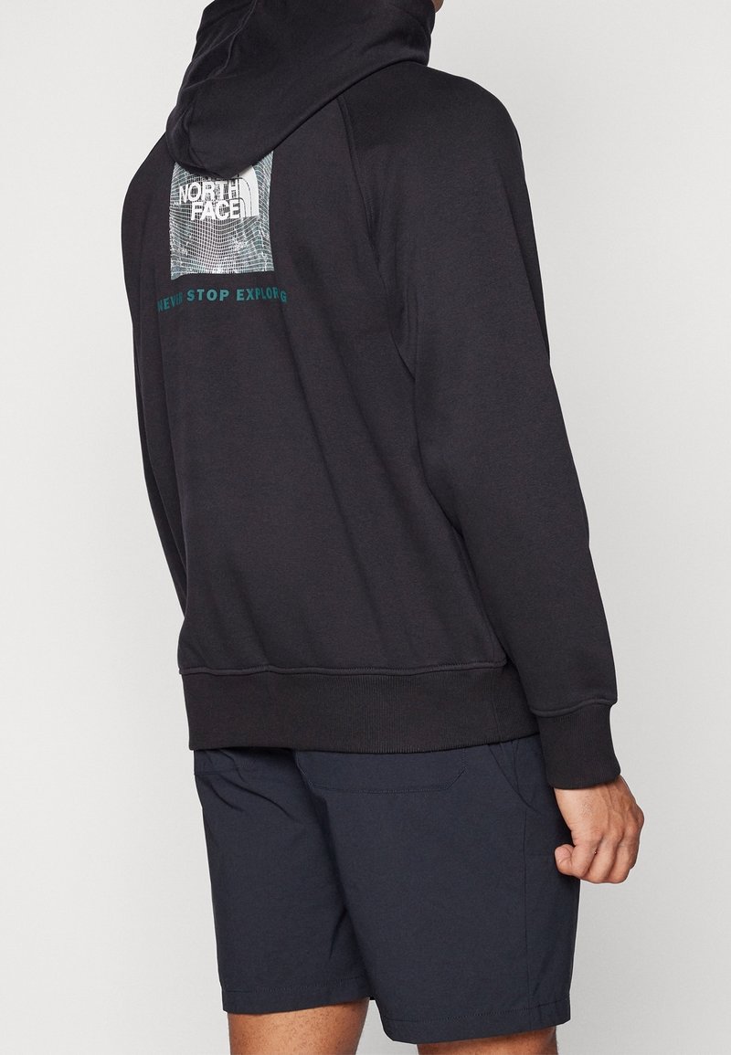 Personne portant un hoodie noir North Face avec logo et texte "Never Stop Exploring" dans le dos, assorti à un short bleu marine foncé.