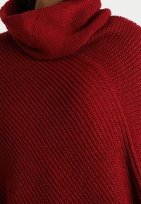 Gros plan sur un pull en maille rouge côtelée avec un col montant épais et replié.