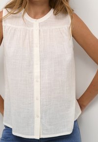 Ærmeløs hvid tekstureret bluse med knapper foran, iført med blå jeans, hænderne hvilende på hofterne mod en hvid baggrund.