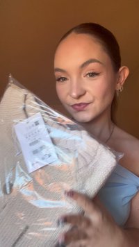 Beżowy sweter robiony na drutach z fakturowanym wzorem, zapakowany w przezroczysty plastik, trzymany w rękach. Widoczna biała metka z danymi produktu.