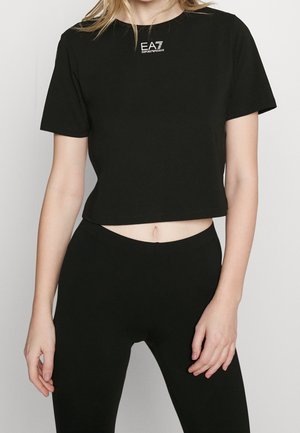 Kobieta w dopasowanym, czarnym, krótkim t-shircie z białym logo "EA7 Emporio Armani" oraz pasujących czarnych legginsach na szarym tle.