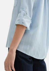 Hellblaue Langarmbluse aus strukturiertem Stoff mit vertikalen weißen Streifen; besitzt hochgerollte Ärmel, die mit Knopflaschen gesichert sind.