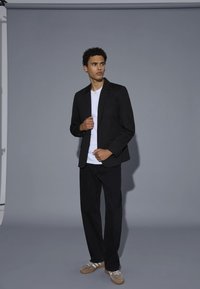 Sacou negru cu un singur nasture și rever de tip notch, cămașă albă dedesubt, asortat cu pantaloni negri și adidași gri.
