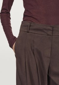 Brązowe, teksturowane spodnie z płaskim pasem i kieszeniami bocznymi, z plisami z przodu, w zestawieniu z bordową bluzką z długim rękawem.