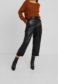 Schwarze Lederhose mit hohem Bund und lockerer Passform, mit umgeschlagenen Säumen. Kombiniert mit einem braunen, gerippten Strickpullover. Schwarze Knöchelstiefel.