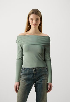 Top verde cu umeri goi și mâneci lungi, cu detalii de material frunțat la gât. Asociat cu blugi albastri decolorati.