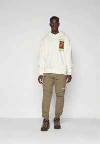 Krēmkrāsas kapuce ar priekšpuses grafiku un ribveida aprocēm; kopā ar haki krāsas šaurām biksēm ar sānu kabatām; izskatu papildina melni pārgājienu zābaki.