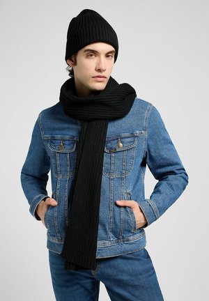 Sort ribbet strikket hue og matchende tørklæde, båret med en blå denimjakke med to frontlommer og guldfarvet hardware, hvilket skaber et lagdelt udseende.