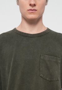 Chemise en coton vert olive à manches longues, avec un col rond et une poche sur la poitrine gauche, présentant une texture côtelée douce et une coupe décontractée.