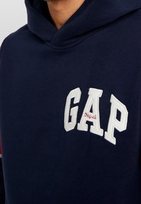 Felpa navy in cotone, con un grande logo "GAP" ricamato in bianco, dal finish texture e un design con cappuccio accogliente.