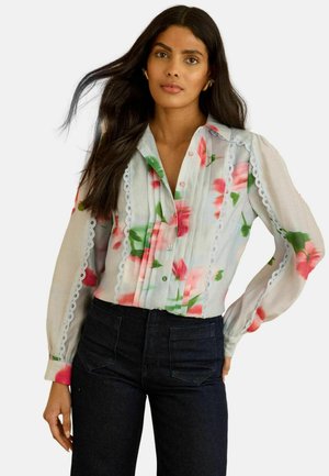 Florale Bluse aus transparentem Stoff mit rosa und grünen Mustern, durchgehender Knopfleiste, Spitzenbesatz an den Seiten und langen Ärmeln. Kombiniert mit dunkler Jeans.