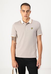 Šedé polo tričko s kontrastním béžovým límcem a lemy rukávů, na hrudi malé logo žluté můry. Černé kalhoty, jednobarevné pozadí.