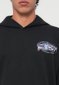 Un hoodie noir en tissu texturé avec un design graphique multicolore sur la poitrine, représentant des éléments abstraits dans des teintes de bleu et de rose.
