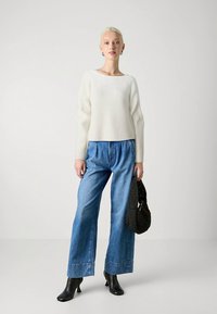 Pulover cu dungi ușoare, blugi largi albaștri, cizme negre cu toc și o geantă neagră țesută. Culori neutre cu siluetă relaxată și texturi variate.