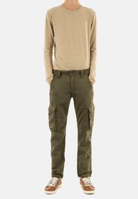 Beige langemouwen shirt, olijfgroene cargobroek met zijzakken en bruine sneakers met witte veters, staand op een effen achtergrond.