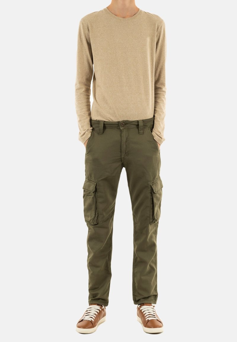 Beige langemouwen shirt, olijfgroene cargobroek met zijzakken en bruine sneakers met witte veters, staand op een effen achtergrond.