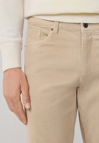 Nahaufnahme einer Hand, die neben beigefarbenen Cordhosen und dem Bund sowie dem Ärmel eines cremefarbenen Rippstrickpullovers ruht.