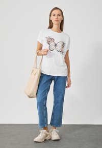 Hvid kortærmet t-shirt med et sommerfugle- og blomstermotiv. Sammen med lyseblå cropped jeans og en beige tote bag. Lyse sneakers.