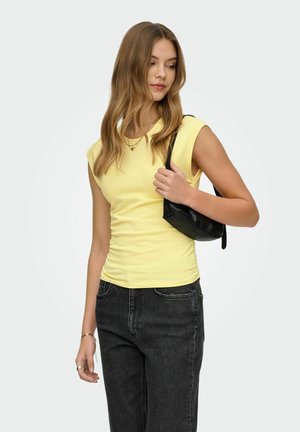 Ung kvinde iført gul ærmeløs top og sorte jeans, der holder en lille sort skuldertaske og kigger mod højre.