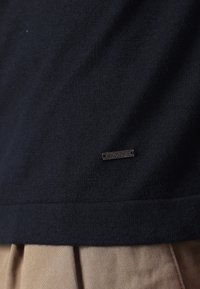 Navy knit sweater featuring a textured finish and a metal logo tag. The hem cuts straight, above a pair of beige pleated trousers.