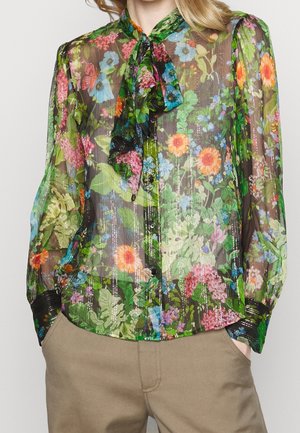 Przezroczysta bluzka z długim rękawem w kolorowy kwiatowy wzór, zapinana na guziki z przodu, z przewiązaną kokardą przy kołnierzyku, zestawiona z beżowymi spodniami i rękami w kieszeniach.