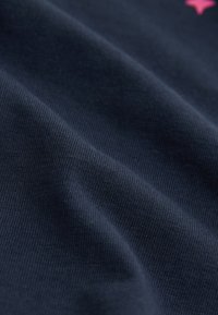 Temno modra tkanina z gladko teksturo, s subtilnim rebrastim vzorcem. Vidne gube kažejo na mehkobo in prožnost, s podrobnostjo roza zvezde.