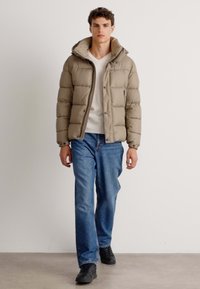 Doudoune marron clair avec capuche, présentant des sections matelassées horizontales et une fermeture à l'avant. Portée sur une chemise blanche et un jean bleu.