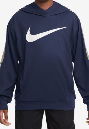 Osoba nosząca granatową bluzę z kapturem z dużym białym logo Nike Swoosh na piersi oraz beżowymi paskami na rękawach.