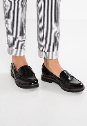 Mocassins en cuir verni noir avec une finition lisse, un petit talon et un détail de sangle décorative, associés à des pantalons rayés retroussés.