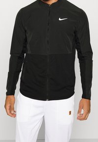Black zippered jacket with a mesh lower section, high collar, and white Nike logo on the upper left. Long sleeves, smooth texture.