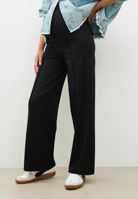 Pantaloni negri cu picioare largi, din material elastic. Asortați cu o jachetă din denim deschis și adidași albi cu accente maro.