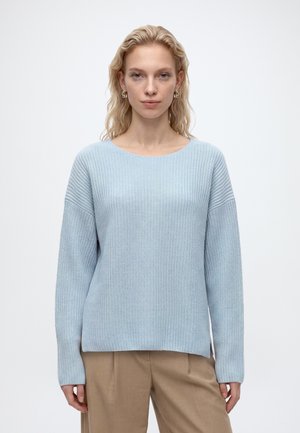 Femme aux cheveux blonds ondulés portant des boucles d'oreilles en or, un pull en tricot bleu clair et un pantalon beige, debout devant un fond blanc uni.