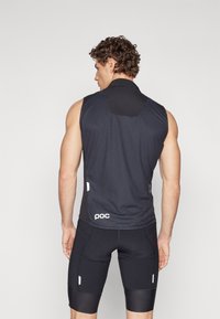 Haut de sport sans manches noir en tissu texturé, avec un col haut et un détail de logo. Porté avec un short noir ajusté et des rayures d'accent.