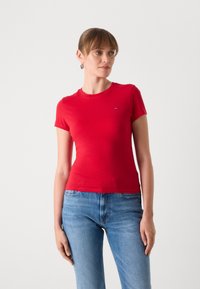 T-shirt rouge à manches courtes en coton, avec un col rond et un petit logo sur la poitrine gauche. Porté avec un jean en denim bleu.