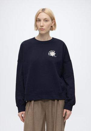Ung kvinde med kort blondt hår iført guldbøjleøreringe, en navy sweatshirt med et hvidt blomstermotiv og beige bukser mod en ensfarvet baggrund.