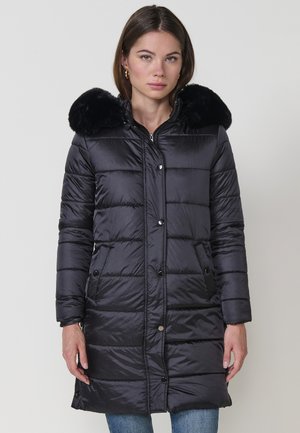 Mujer joven vestida con un abrigo de invierno largo y negro acolchado, con un cuello de piel sintética negro, de pie contra un fondo claro y liso.