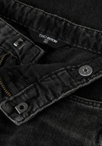 Pantalons en denim noir avec une fermeture éclair et un bouton en métal. Présente une étiquette portant la mention "TWO SOON" et des détails de couture visibles autour de la taille.