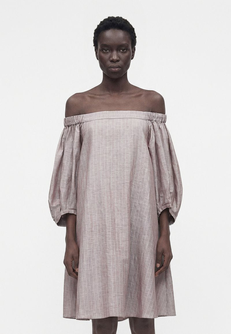 Γυναίκα με φαρδιά, ανοιχτό ροζ φόρεμα εκτός ώμων με κάθετες λευκές ρίγες και φουσκωτά μανίκια τριών τετάρτων.