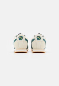 Scarpe sportive con una tomaia crema, dettagli in teal e una finitura in suede testurizzato. Include un logo dorato e una suola in gomma con una tonalità arancione.