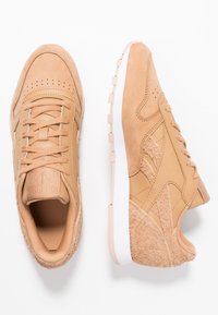 Bruna sneakers med mocka- och läderpaneler, texturerad häl, vit sula och tonade snörningar, visade från ovan- och sidovy mot vit bakgrund.