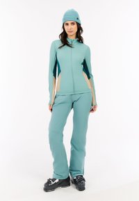 Žena oblečená v tyrkysovom zimnom športe s béžovými bočnými panelmi, s rebrovanou čiapkou a čiernymi lyžiarskymi topánkami, stojí na bielom pozadí.