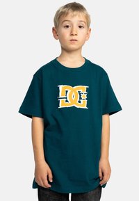 Teal bomulds T-shirt med et hvidt og orange DC-logo trykt på forsiden. Løs pasform, korte ærmer, rund halsudskæring. Ingen synlige mønstre.