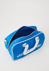 Blå bag med hvite detaljer, glidelås-lukking, doble håndtak, og teksturert "adidas"-logo mønster på overflaten. Innsiden er foret i blå.